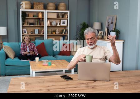 Glücklicher alter Mann huzband Fernarbeit oder Fernunterricht online vom Home Office aus über Laptop Computer Videoanrufe während seine Frau im Büro sitzt Stockfoto