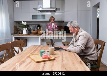 Glücklicher älterer Mann Ehemann, der aus der Ferne arbeitet oder online Fernunterricht mit einem Laptop vom Heimbüro aus lernt, während ihre ältere Frau Frühstück kocht Stockfoto