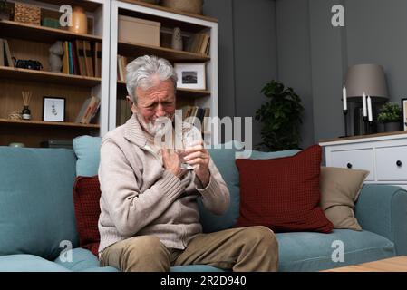 Älterer Mann bekommt Brustschmerzen im Wohnzimmer, Herzinfarkt hält seine Hand auf der Brust und sitzt zu Hause auf dem Sofa. Gesundheitsfürsorge für ältere Menschen Stockfoto