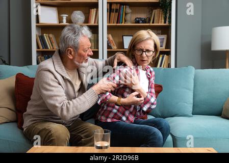 Porträt eines älteren Ehepaars im Wohnzimmer, eine reife Frau mit Schmerzen in der Schulter. Oberarmschmerzen, Nacken- und Schulterschmerzen der Frau und Rückenverletzung Stockfoto