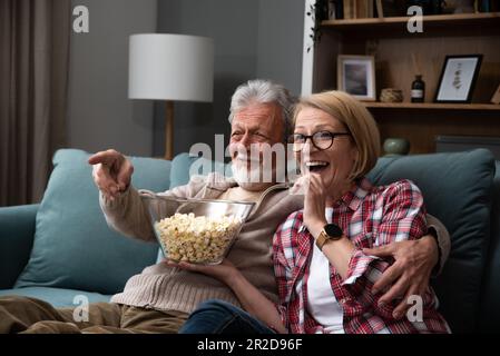 Lachendes älteres Paar, Mann und Frau, die fernsehen, Comedy-Show oder Film schauen und Popcorn-Snack essen, zu Hause auf einer gemütlichen Couch sitzen, ältere Familie, Mann und Stockfoto