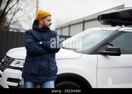 Der Fahrer trägt bei kaltem Wetter eine Jacke und einen gelben Hut gegen seinen amerikanischen SUV mit Dachgepäckträger. Stockfoto