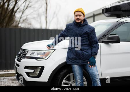 Der Fahrer trägt bei kaltem Wetter eine Jacke und einen gelben Hut gegen seinen amerikanischen SUV mit Dachgepäckträger. Stockfoto