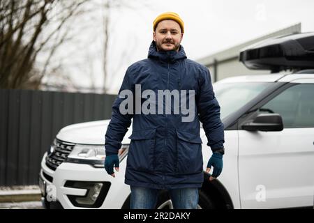 Der Fahrer trägt bei kaltem Wetter eine Jacke und einen gelben Hut gegen seinen amerikanischen SUV mit Dachgepäckträger. Stockfoto