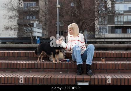 Junge, wunderschöne Hipsterfrau sitzt auf der Treppe mit ihrem Pflegehund, den sie aus einem Tierheim adoptiert hat, und gibt ihm Liebe und Zärtlichkeit Stockfoto