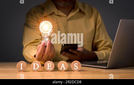 Ein Geschäftsmann, der eine Glühbirne auf einem Holzblock hält, mit Textideen für Unternehmen, ein neues Ideenkonzept mit Innovation und Inspiration, Innovation Stockfoto