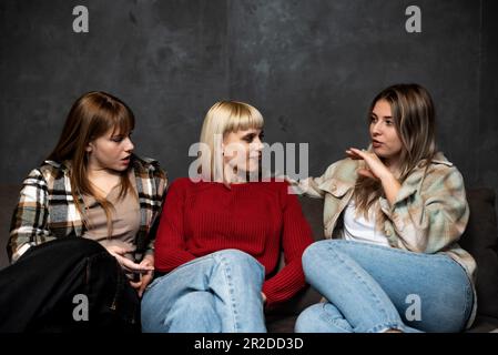 Drei junge Freundinnen sitzen zu Hause auf dem Sofa und klatschen über andere Frauen und Menschen, reden über ihr Leben und machen Witze über eac Stockfoto