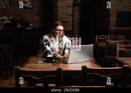 Junge Geschäftsfrau, die Inhaber oder Manager einer gemütlichen Bar und Cafeteria ist, sitzt nach Ende der Arbeitszeit und kontrolliert Einnahmen und Umsatz auf einem Laptop c Stockfoto