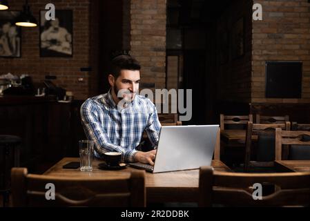 Ein junger Geschäftsmann, der Inhaber oder Manager einer gemütlichen Bar und Cafeteria ist, sitzt nach Ende der Arbeitszeit und kontrolliert die Einnahmen und den Umsatz über ein Notebook Stockfoto