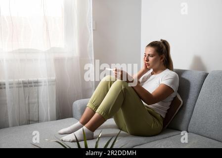 Junge nervöse Frau sitzt zu Hause und hält einen Schwangerschaftstest ab und wartet ungeduldig auf das Testergebnis. Vielleicht schwanger, vielleicht nicht mit Concept Stockfoto