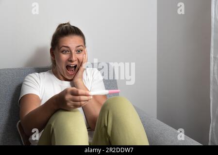 Schließlich schwanger. Attraktive junge Frauen bei Schwangerschaft Test suchen und lächelnd, während sie auf dem Sofa zu Hause sitzen Stockfoto