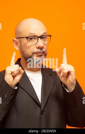 Ein stilvoller Mann in einer Anzugjacke, mit Bart, Glatze und Brille vor orangefarbenem Hintergrund Stockfoto