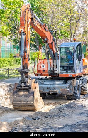 Ein großer Mobilbagger steht in einem Graben, den er an einem hellen, sonnigen Frühlingstag mitten auf einer Straße gegraben hat. Vertikales Bild. Stockfoto