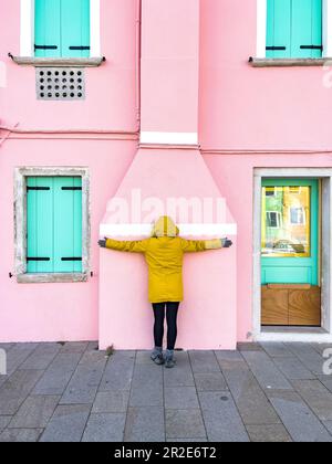 Nicht wiedererkennbare Person in gelber Regenjacke auf rosa Wandhintergrund. Rückblick auf Touristen, die auf einem wunderschönen rosa Haus mit türkisfarbenen Fensterläden posieren Stockfoto