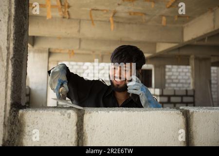 Ein glücklicher bauarbeiter baut eine Zementmauer, während er telefoniert. Stockfoto