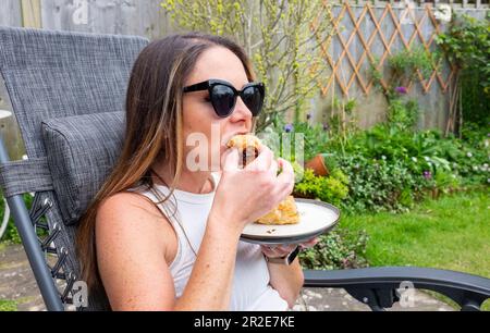 Junge Frau in ihren 30s Jahren isst eine Wurstrolle zum Mitnehmen in einem kleinen Stadtgarten im Frühlingssonnenschein Credit Simon Dack Stockfoto
