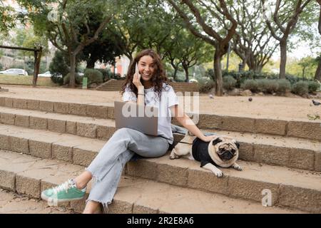 Positiver und lockiger junger Freiberufler, der auf einem Smartphone in der Nähe eines Laptops spricht, in die Kamera schaut und den Hund streichelt, während er auf der Treppe im Park in Barcel sitzt Stockfoto