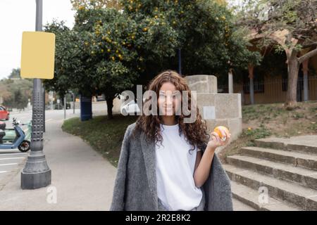 Lächelnde junge, lockige Frau in warmer Jacke, die frisches Orange hält und in die Kamera schaut, während sie auf einer verschwommenen urbanen Straße im Hintergrund in Barcel steht Stockfoto