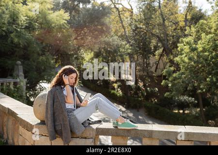 Junger, lockiger Freiberufler in warmer Jacke mit Smartphone und Ohrhörern, während er seinen Laptop in der Hand hält und im verschwommenen Sommerpark in der Bar auf einem Steinzaun sitzt Stockfoto