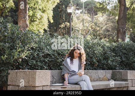 Junger, lockiger Freiberufler in Freizeitkleidung, der sein Smartphone benutzt und auf dem Notebook schreibt, während er neben dem Laptop auf einer Steinbank und grünen Büschen in Par sitzt Stockfoto