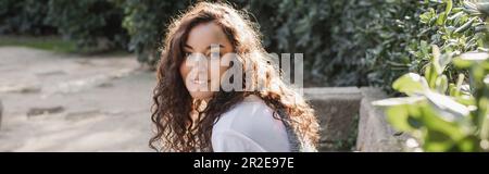 Lockige und junge brünette Frau in einem T-Shirt, die vor der Kamera lächelt, während sie auf einer Steinbank sitzt und sich im Park in Barcelona, Spanien, in der Nähe der grünen Büsche entspannt Stockfoto