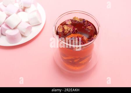 Gesunde Kräuterblütenblätter, Knospen Tee in doppelwandigen Glasbecher, Tasse. Rosafarbener weißer Marshmallow-Teller. Rosafarbener Hintergrund. Getrocknete bunte Blütenblätter Stockfoto