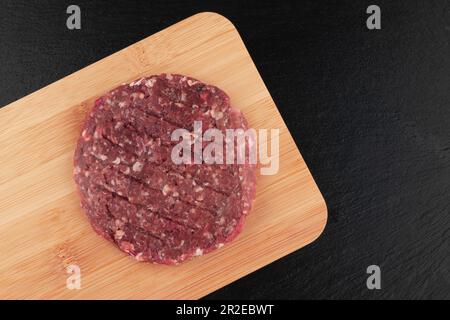 Rohe Hamburger-Bratlinge auf Holzschneidebrett auf schwarzem Hintergrund Stockfoto