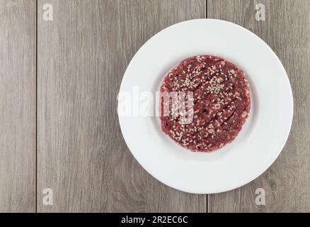Rohes Hamburger-Steak mit Sesamsamen auf weißem Teller auf Holzhintergrund. Stockfoto