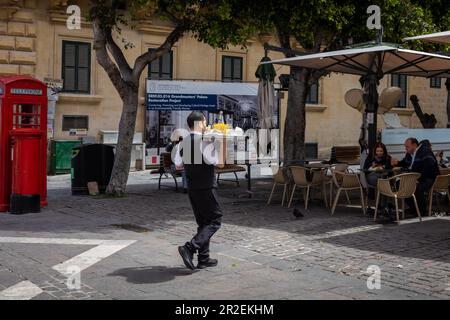 Valletta, Malta - 16. April 2023: Ein Kellner, der ein Serviertablett in einem Café im Freien am Platz der Republik mit sich führt. Stockfoto