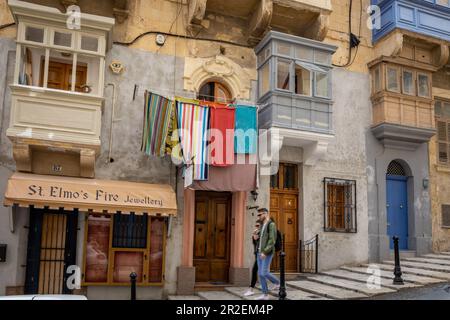 Valletta, Malta - 16. April 2023: Touristen gehen die Straße entlang mit traditionellen maltesischen Balkonen und farbenfroher Wäscherei. Stockfoto