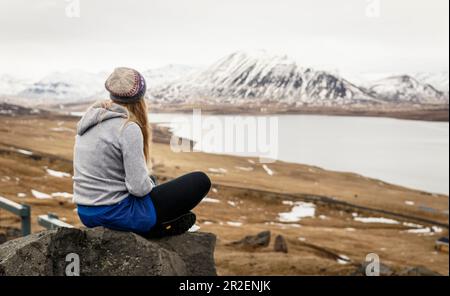 Junge Frau sitzt auf Stein und blickt auf leicht verschneite isländische Landschaft, Island Stockfoto