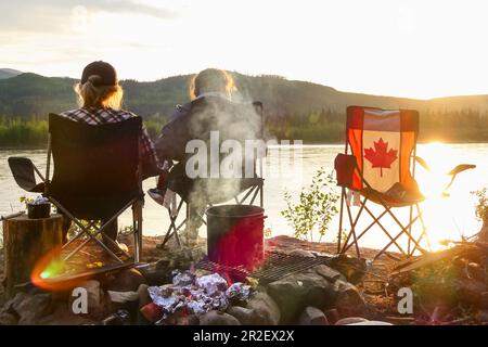 Zwei Mädchen sitzen bei Sonnenuntergang am Feuer und genießen den Blick auf den Yukon River. Kanada, Whitehorse, Yukon Stockfoto