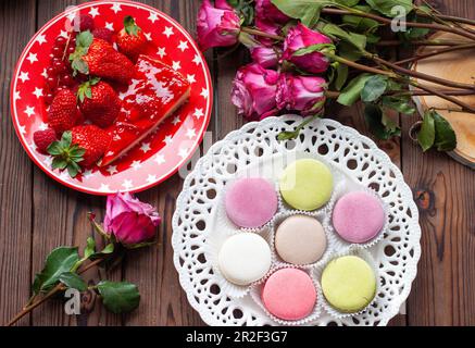 Eine Scheibe Erdbeerkuchen, Makronen- und Rosenblume auf einem roten Teller, von oben auf einen Holztisch gelegt. Wunderschönes Urlaubsfrühstück. Flach liegend. Stockfoto