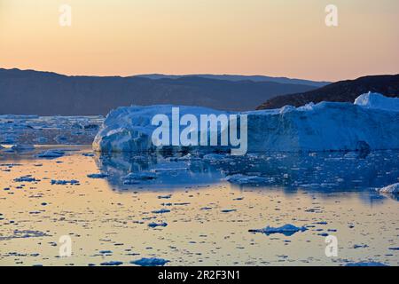 Fjord auf dem Eqi-Gletscher in Westgrönland; Eisberge und Eisschollen im ruhigen Wasser; Berge im Hintergrund; Abendsonne, Himmel und Wasseroberfläche Stockfoto