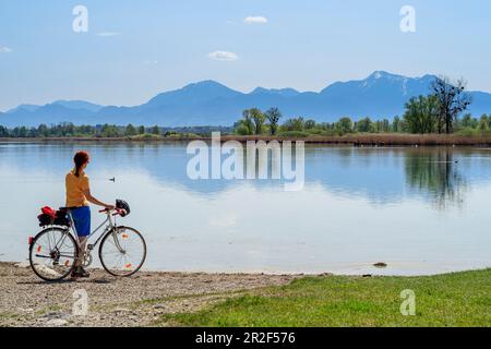 Die Radfahrerin steht am Ufer und schaut auf Chiemsee, Hochfelln und Hochgern im Hintergrund, Chiemseeradweg, Chiemgau, Oberbayern, Bayern, G Stockfoto