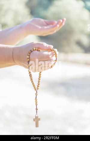 Frauenhände halten einen christlichen Rosenkranz mit einem Kreuz, beten in der Dämmerung auf einem Feld Stockfoto