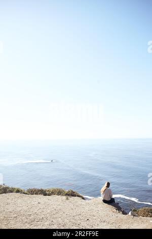 Eine junge Frau sitzt auf einer Klippe und blickt auf den Pazifik am Rande des Highway 1, Big Sur State Park, Kalifornien, USA. Stockfoto