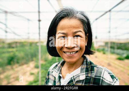 Glückliche Frau aus Südostasien, die in der Landwirtschaft im Gewächshaus vor der Kamera lächelt – „Farm People Lifestyle Concept“ Stockfoto