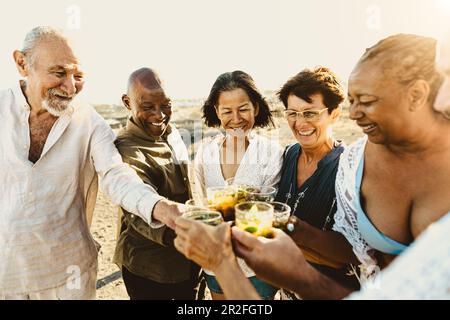 Fröhliche, multirassische Seniorenfreunde, die Spaß beim Trinken und Toasten von Mojitos am Strand während des Sonnenuntergangs haben - ältere Leute genießen Sommerferien Stockfoto