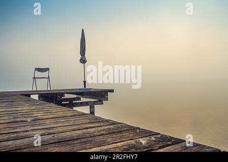 Anlegestelle mit gefalteten Sonnenschirmen bei nebeligem Sonnenaufgang am Starnberger See, Seeshaupt, Bayern, Deutschland Stockfoto