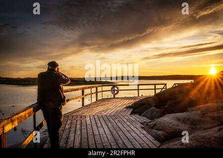 Ein Mann beobachtet den Sonnenuntergang auf einem Steg in den Inselgruppen Ellös, Orust, Bohuslän, Schweden Stockfoto