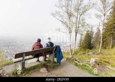 Bergen, Norwegen - Mai 2023: Ein älteres Paar in warmer Kleidung sitzt auf einer Bank auf der Fløyen und genießt den Blick über Bergen, eine Stadt, die zum UNESCO-Weltkulturerbe gehört. Stockfoto