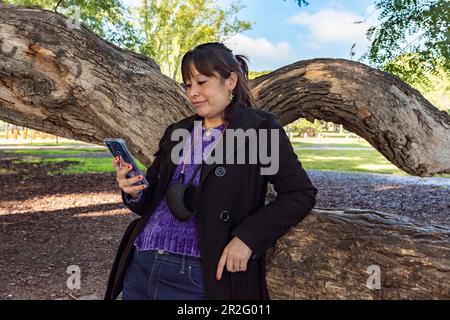 Eine Frau liest eine SMS auf ihrem Handy, während sie sich an einen Baum im Park lehnt Stockfoto