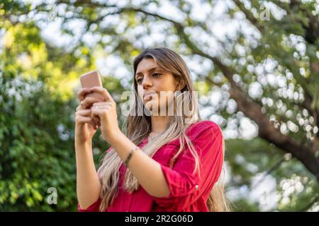 Low-Angle-Betrachtung einer jungen blonden Frau, die ihr Smartphone im Freien benutzt. Modernes Technologiekonzept Stockfoto