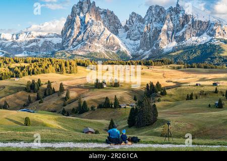 Vater und Sohn genießen den Sonnenuntergang mit Blick auf das schneebedeckte Langkofel und Plattkofel auf der Alpe di Siusi in Südtirol, Italien Stockfoto