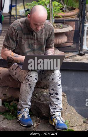 Ein Mann mit tätowierten Armen arbeitet auf einem Laptop im Heimbüro in Garten, Baden-Württemberg, Deutschland Stockfoto