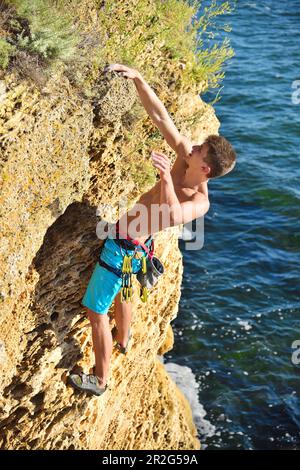 Junger Kletterer, der an einer Klippe auf einem Meereshintergrund hängt Stockfoto