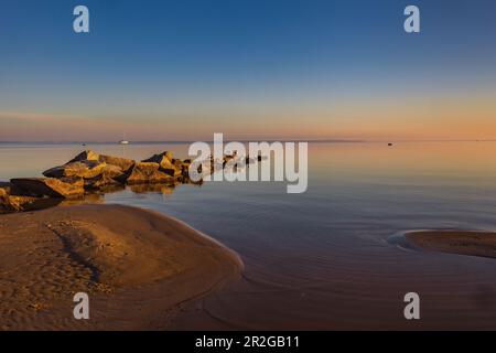 Ostseeküste mit Wellenbrecher bei Sonnenaufgang, im Hintergrund können Sie ein Segelboot sehen. Szczecin Lagoon, Ostsee, Mecklenburg-Vorpommern, Ge Stockfoto