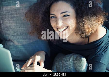 Eine strahlende junge Frau mit lockigem Haar kichert heftig, während sie mit ihrem Computer interagiert. Sie könnte über ein lustiges Meme lachen, ein lustiges V gucken Stockfoto