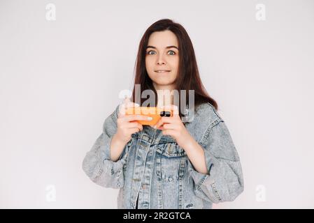 Unentschlossen und unsicher: Junge Frau in Denim-Jacke spielt Smartphone-Spiel, isoliert auf grauem Hintergrund. Betonung von Unsicherheit und Zweifeln Stockfoto